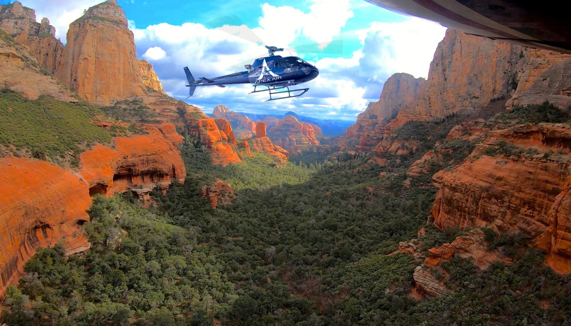 Helicopter flying over a red rock valley surrounded by large red rock formations.