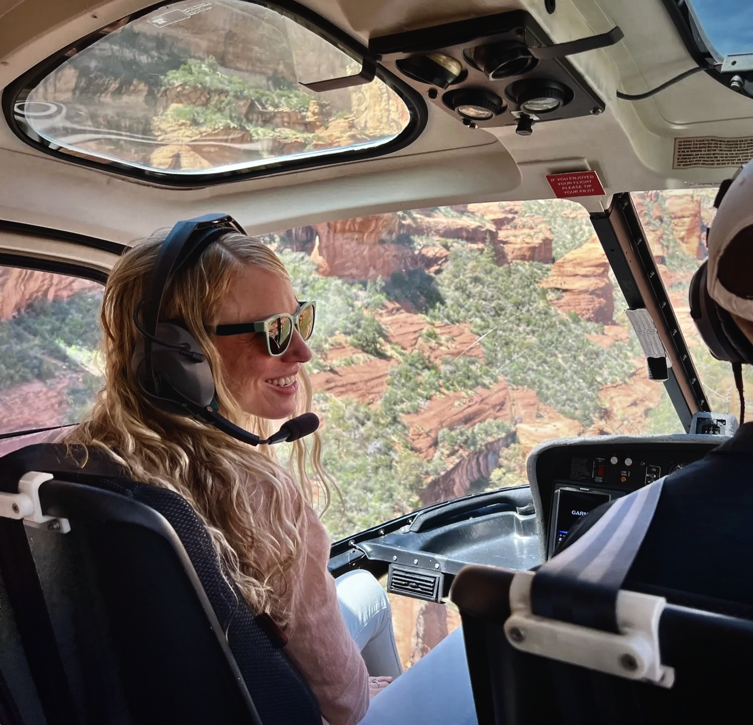 Woman with headset smiling inside helicopter, scenic canyon view outside window.
