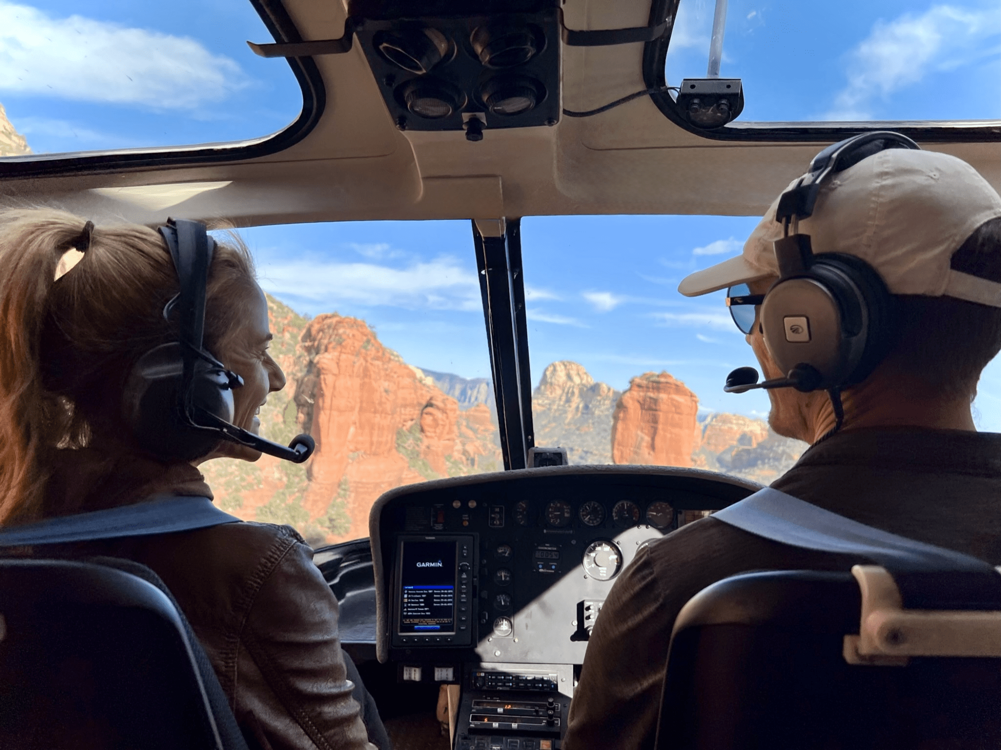 Two people with headsets in a helicopter cockpit view of red rock formations.