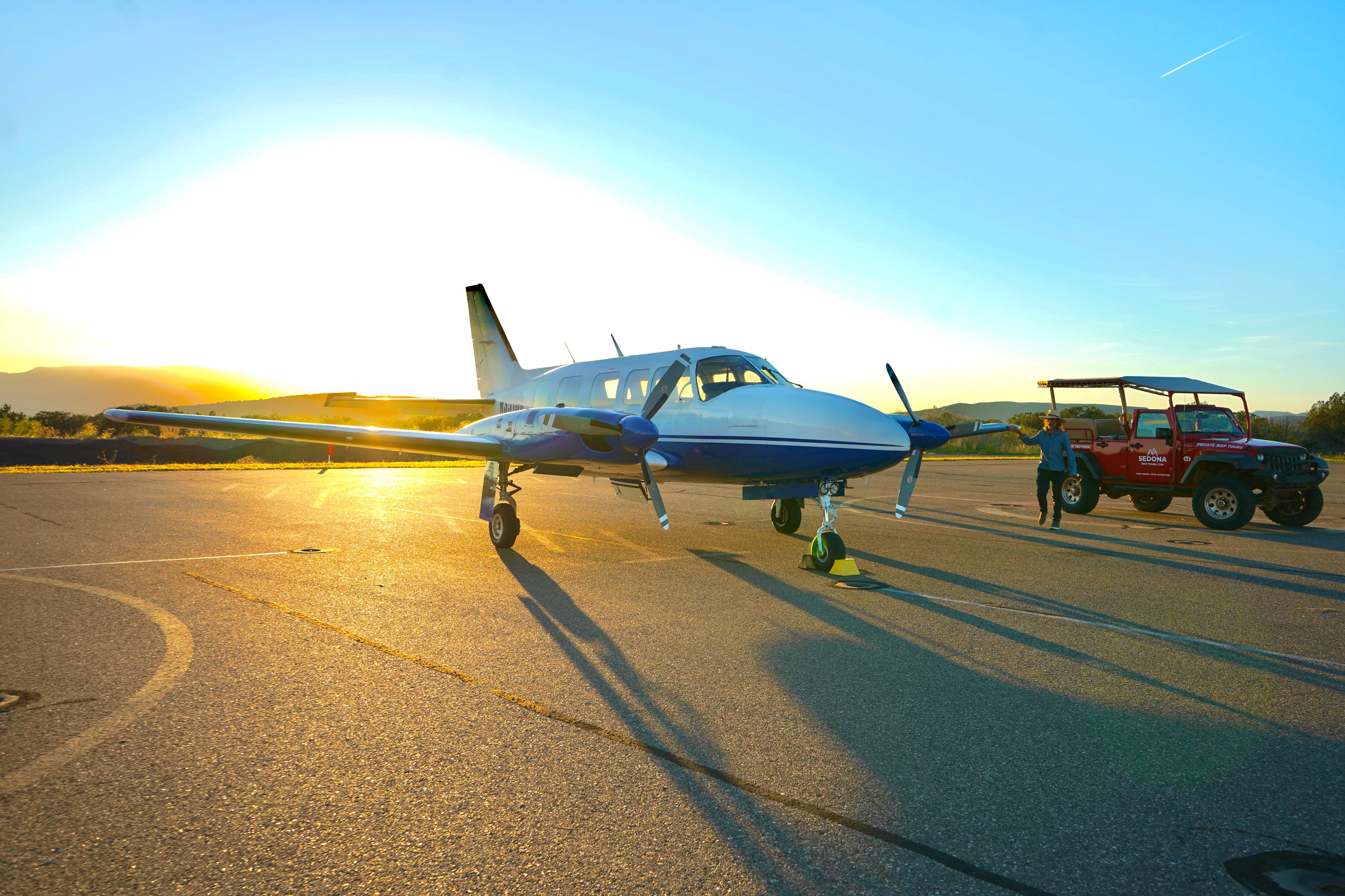Small plane on runway at sunset with man and red jeep nearby.