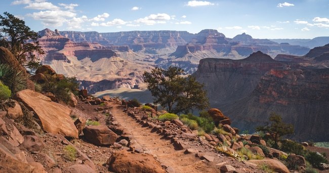 Trail with trees and red rock formations in the Grand Canyon under a blue sky with clouds.