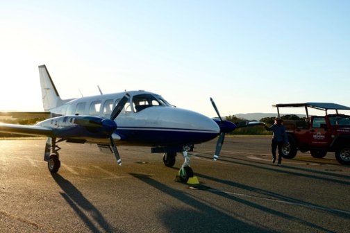 Small aircraft on tarmac with a red off-road vehicle and person in the background at sunset.