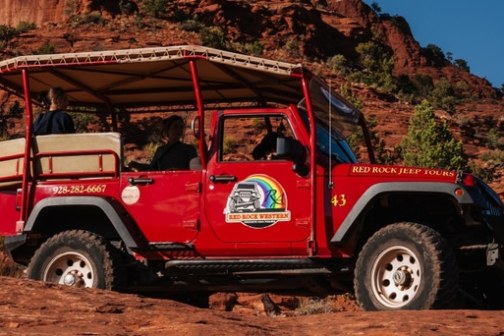 Red jeep with open roof on rocky terrain, passengers visible, desert landscape in background.