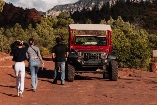 Three people walk towards a red jeep on a rocky, forested landscape.