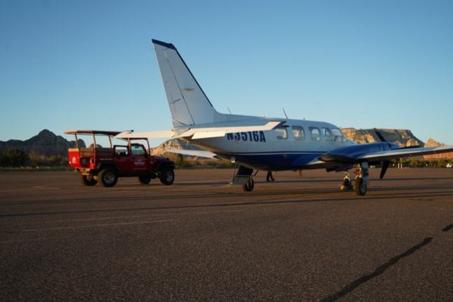 Small airplane and red jeep parked on tarmac with scenic mountains in the background.