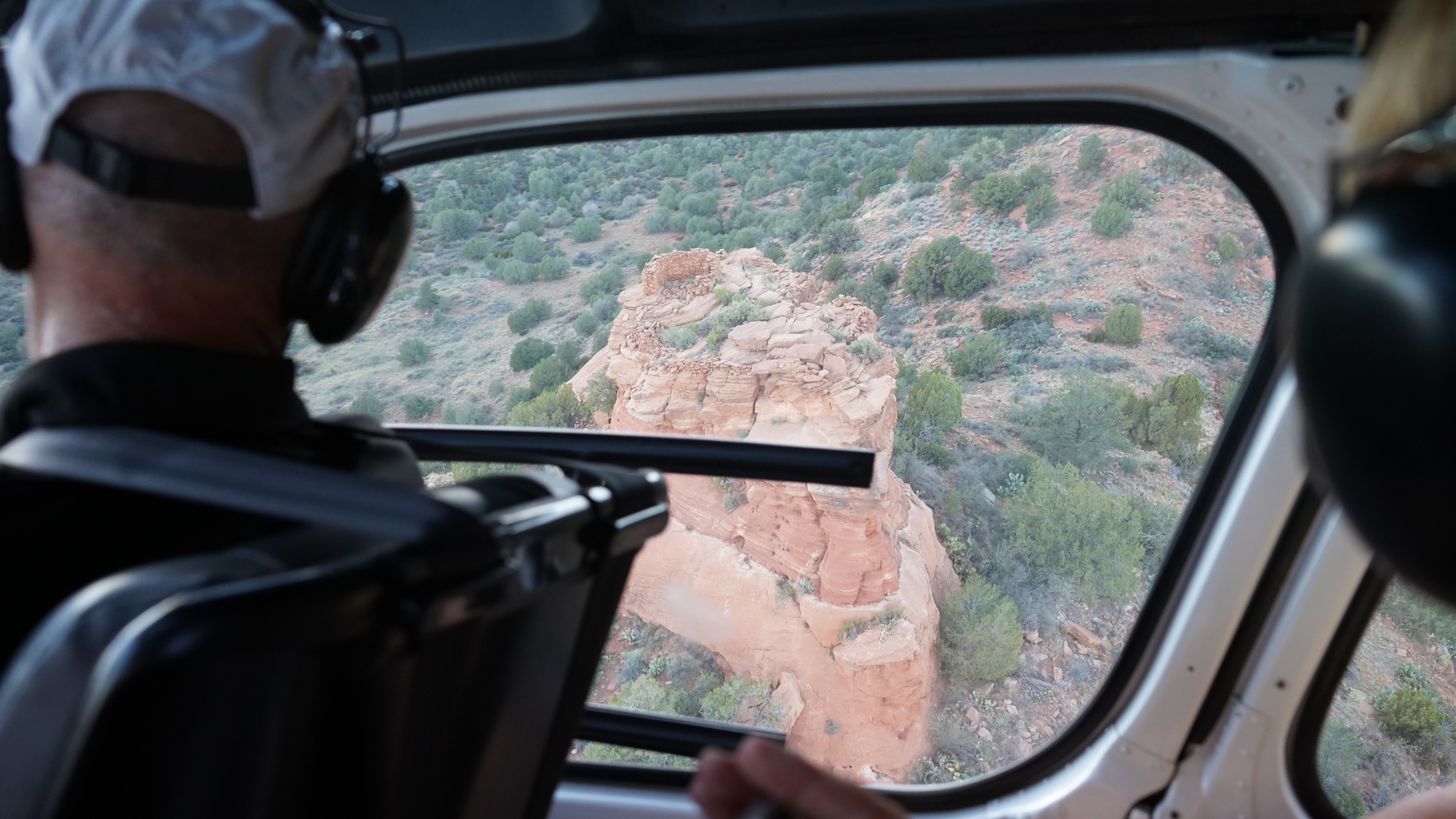View from helicopter window showing red rock formation and green shrubs below.