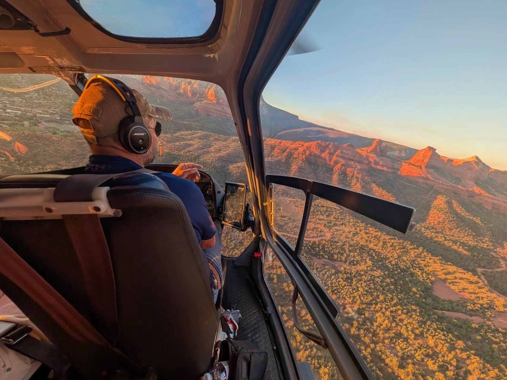 Helicopter cockpit with pilot flying over sunlit red rock formations and forests.