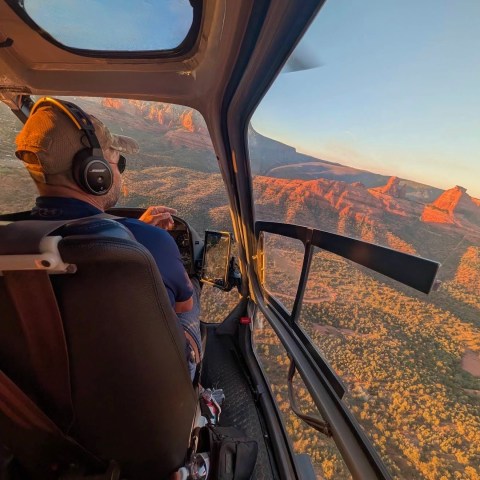 Helicopter cockpit with pilot flying over sunlit red rock formations and forests.